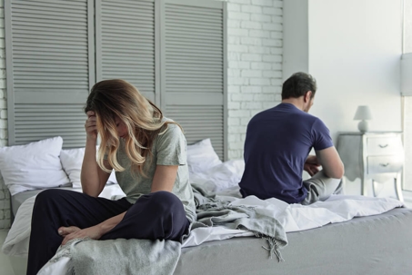 Couple sitting on opposite sides of a bed with their backs to one another.